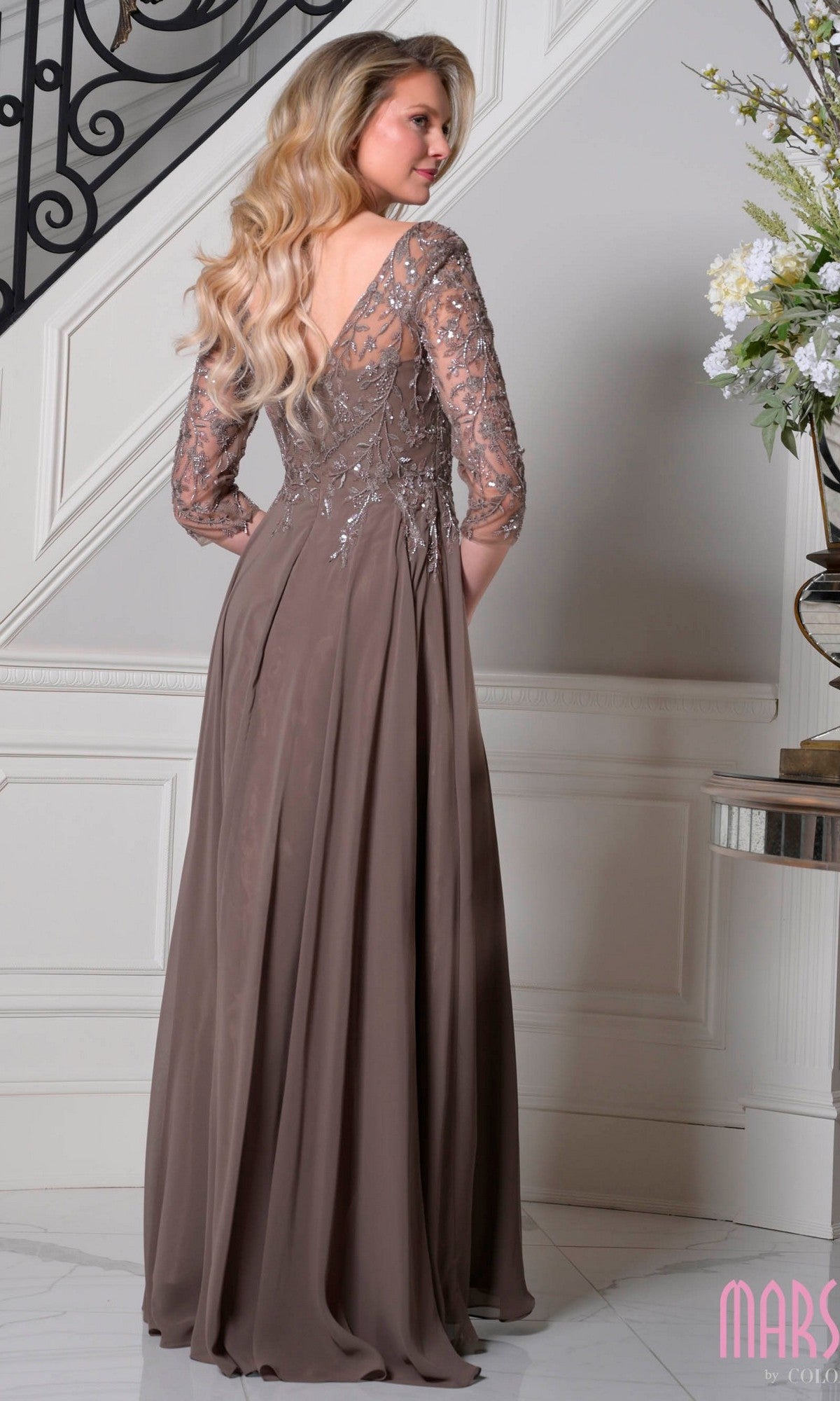 A woman with long blonde hair stands indoors by a staircase wearing the Marsoni M340SL, a modest taupe long formal dress with an A-line chiffon skirt, beaded-embroidered bodice, and sheer embellished back and sleeves.